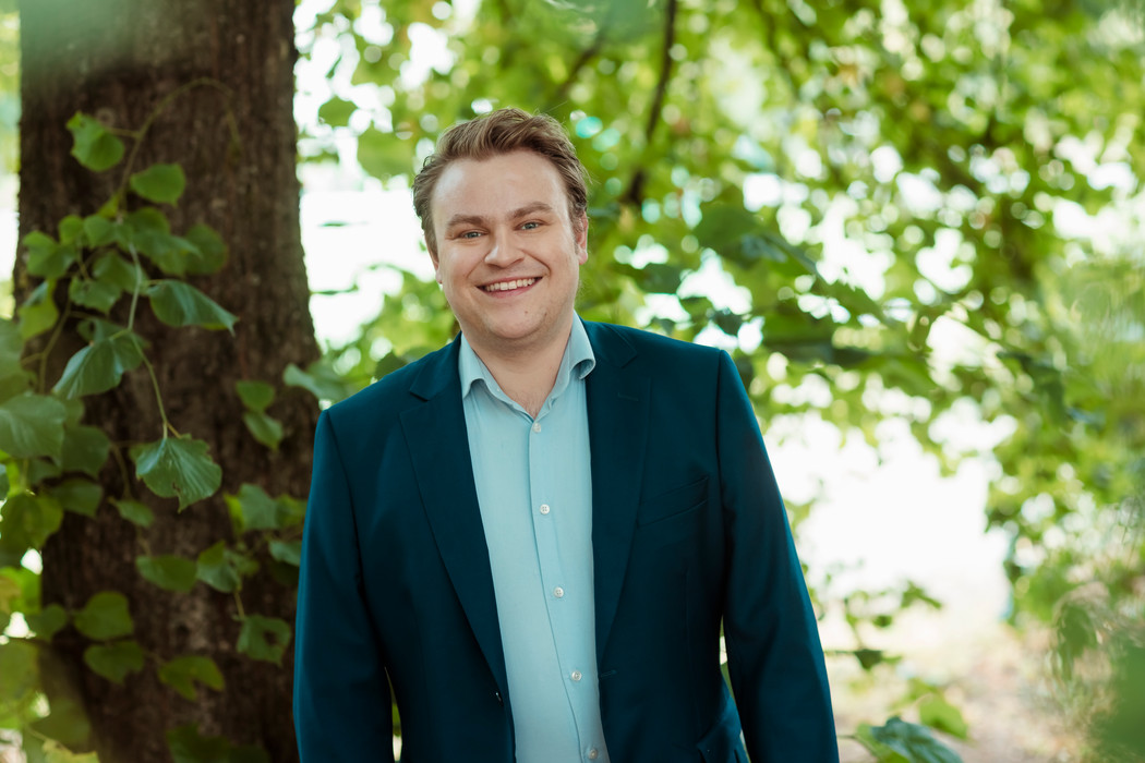 Daniel Naper med blondt hår iført en mørk dress og lys skjorte står med armene ned langs siden og smiler mot kameraet. Foto
