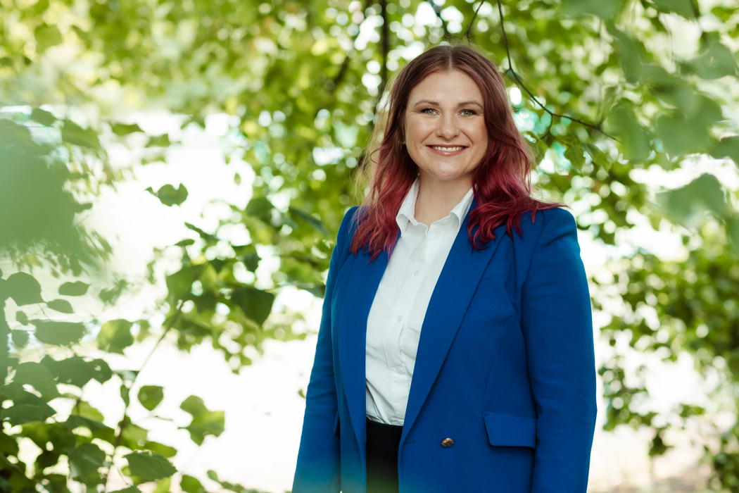 Hanne Reese Holm med rødt hår iført en lys dress og hvit skjorte står med armene i ned langs siden og smiler mot kameraet. Foto.
