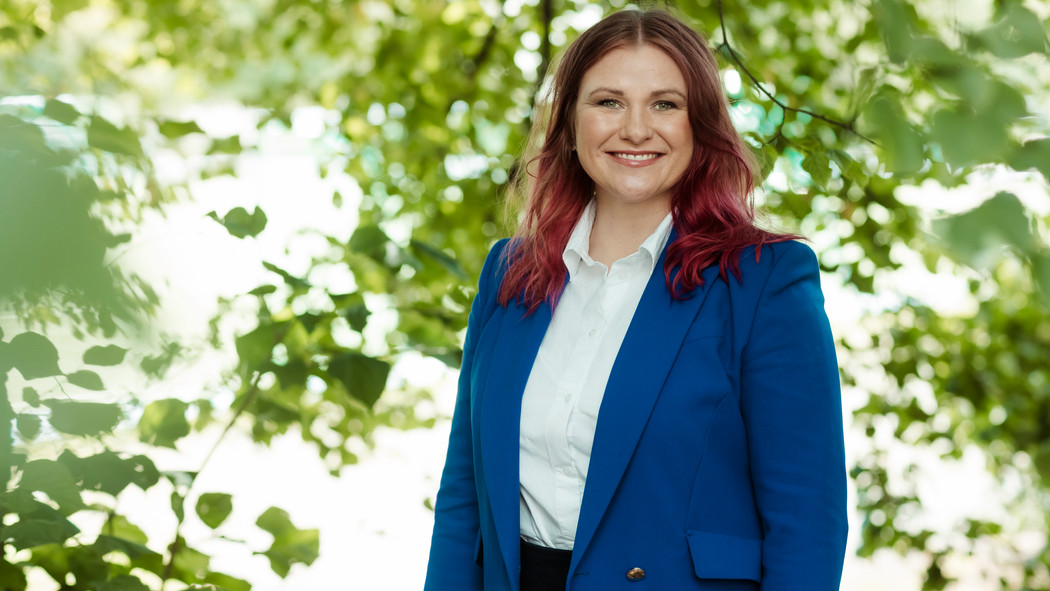 Hanne Reese Holm med rødt hår iført en lys dress og hvit skjorte står med armene i ned langs siden og smiler mot kameraet. Foto.