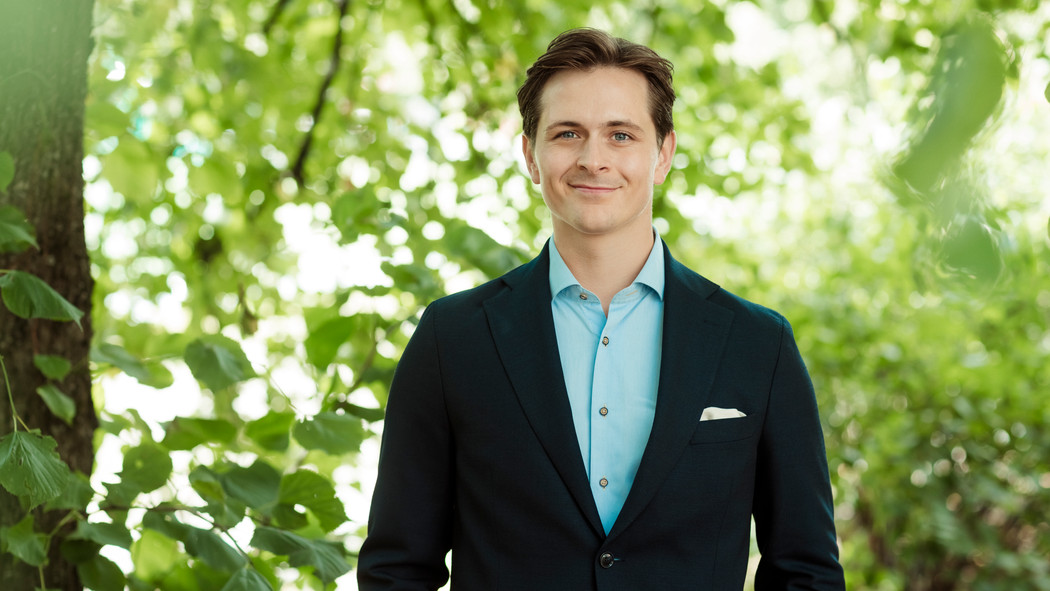 Njaal Søstumoen med kort hår iført en mørk dress og lys skjorte står med armene ned langs siden og smiler mot kameraet i et kontor. Foto.