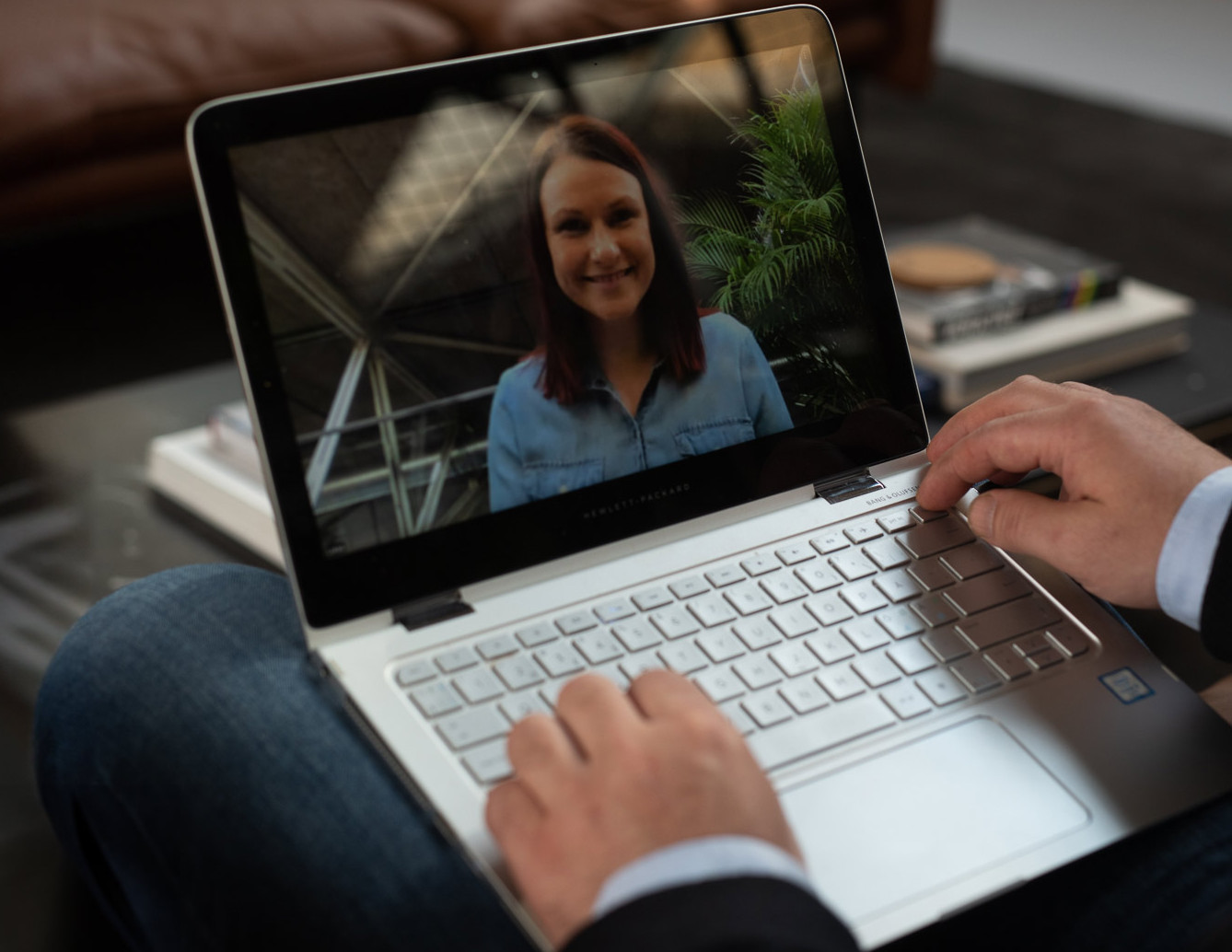 En person i dress sitter med en laptop i fanget, og på skjermen er en smilende kvinne synlig under en videosamtale. Bakgrunnen er et moderne kontor med en brun sofa og bøker på et bord. Foto.